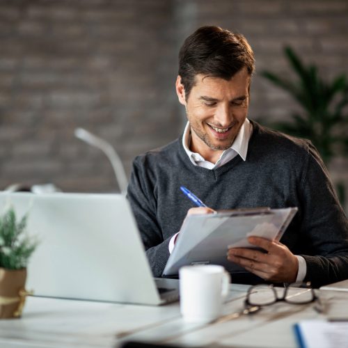 Smiling entrepreneur writing business report while working on laptop in the office.