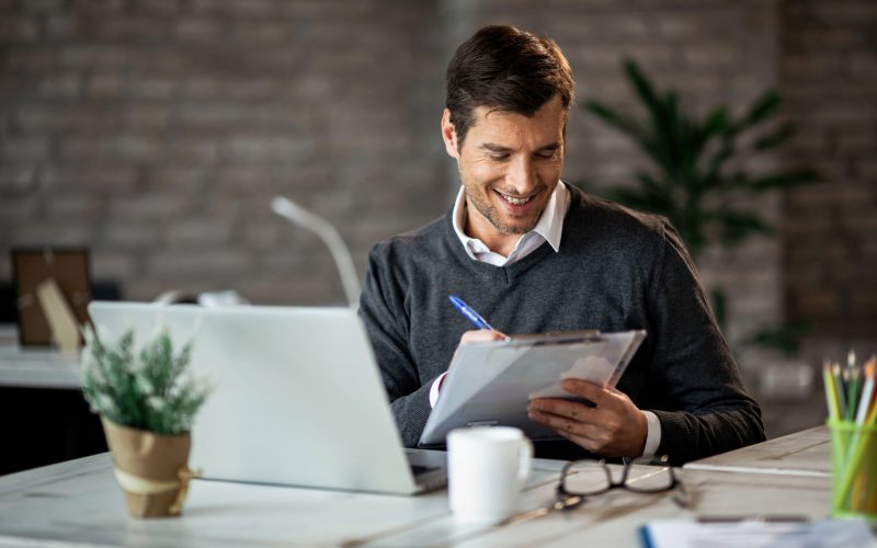 Smiling entrepreneur writing business report while working on laptop in the office.